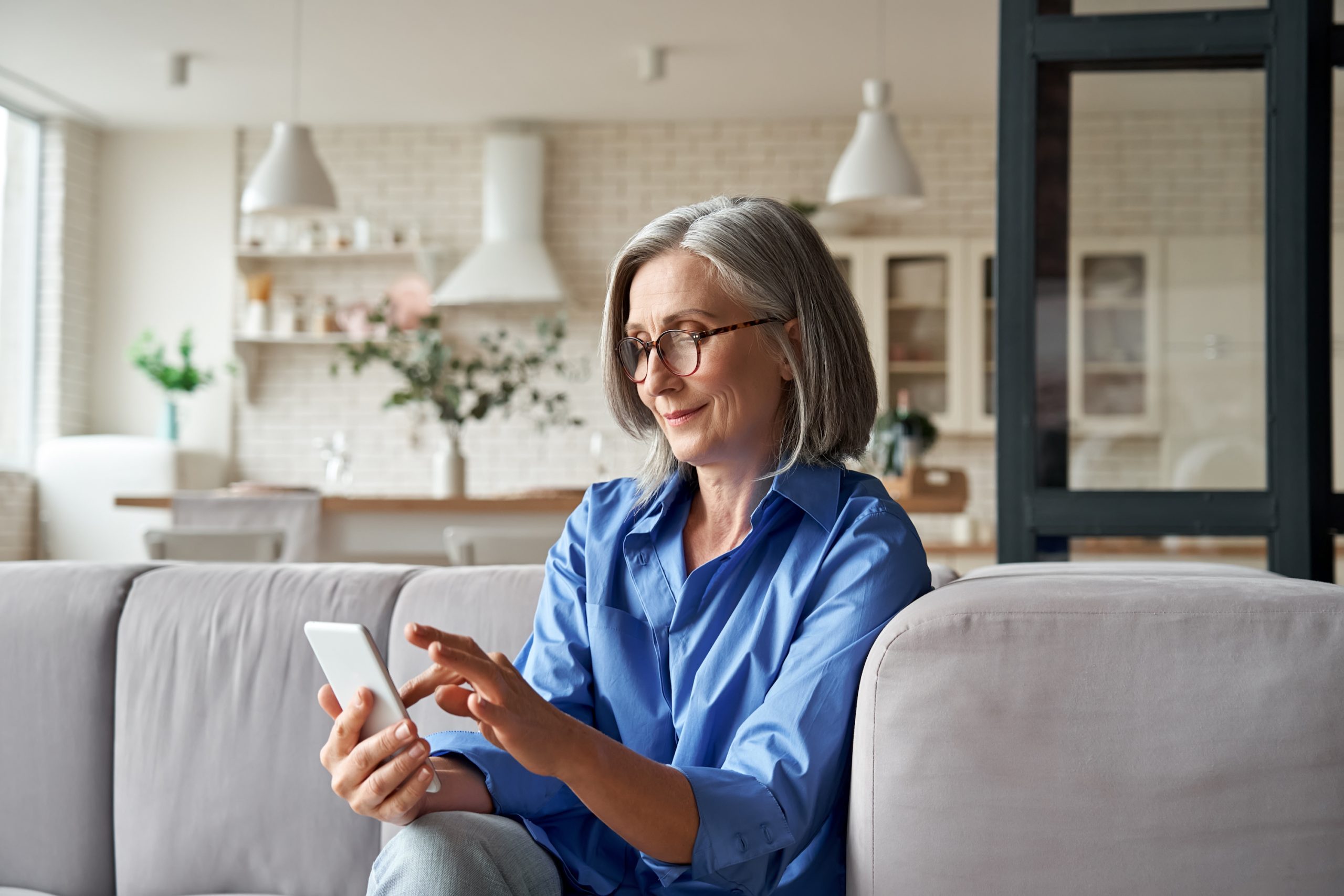 Mature old senior woman using smartphone sitting on sofa at home text messaging